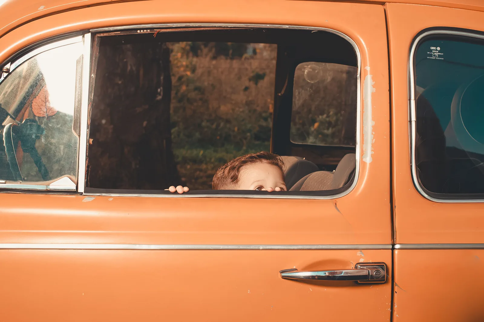 A young child peeking out the window of a vintage orange car