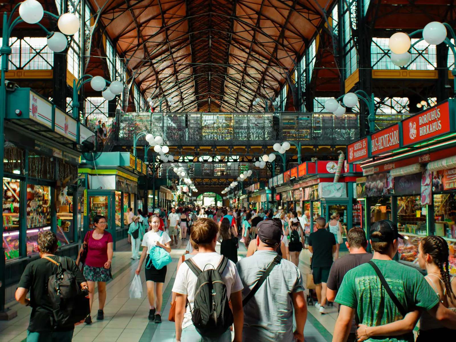 Shoppers walking under the iron roof and hanging lamps inside the Great Market Hall in Budapest
