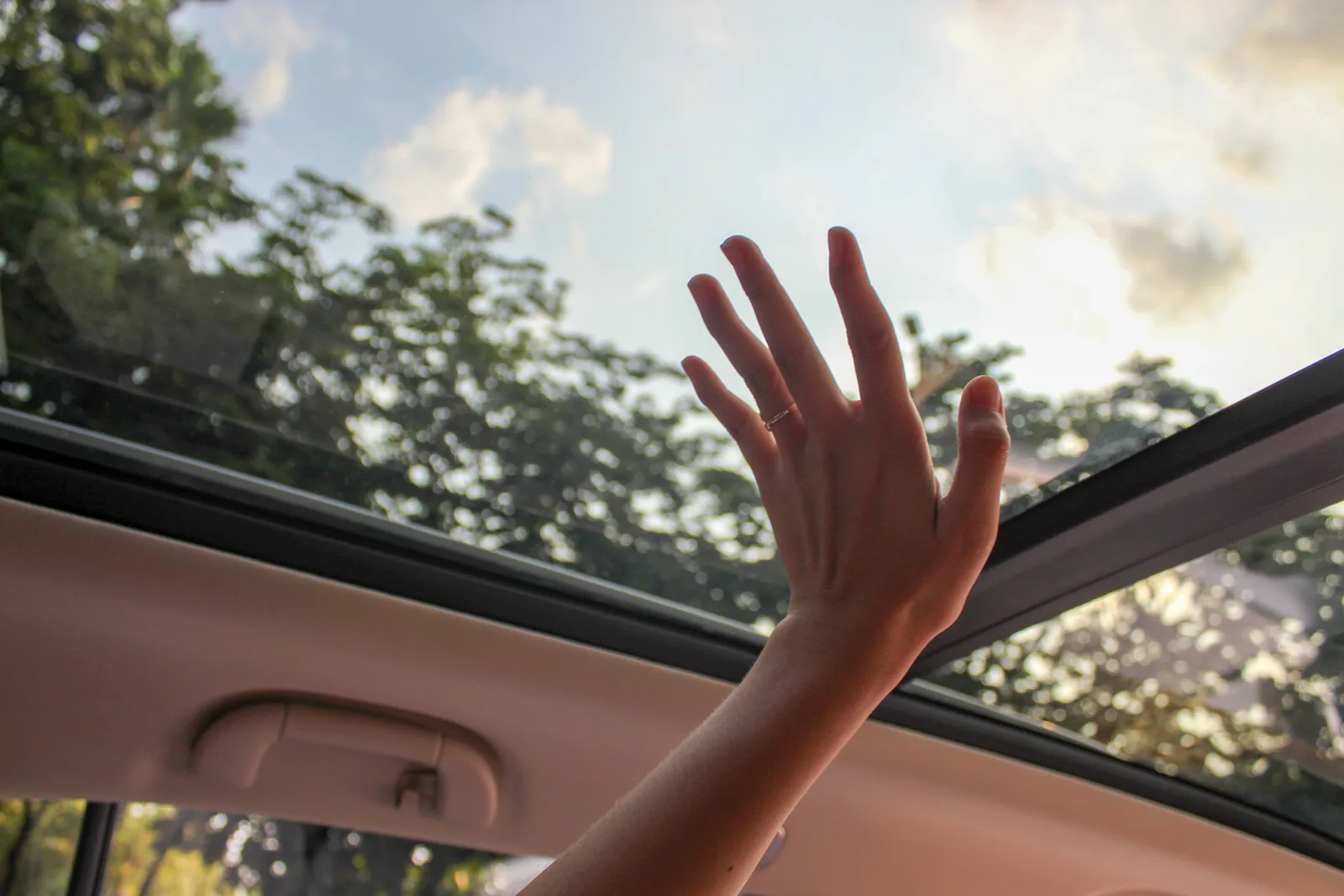 A hand reaching up through an open car sunroof toward the sky