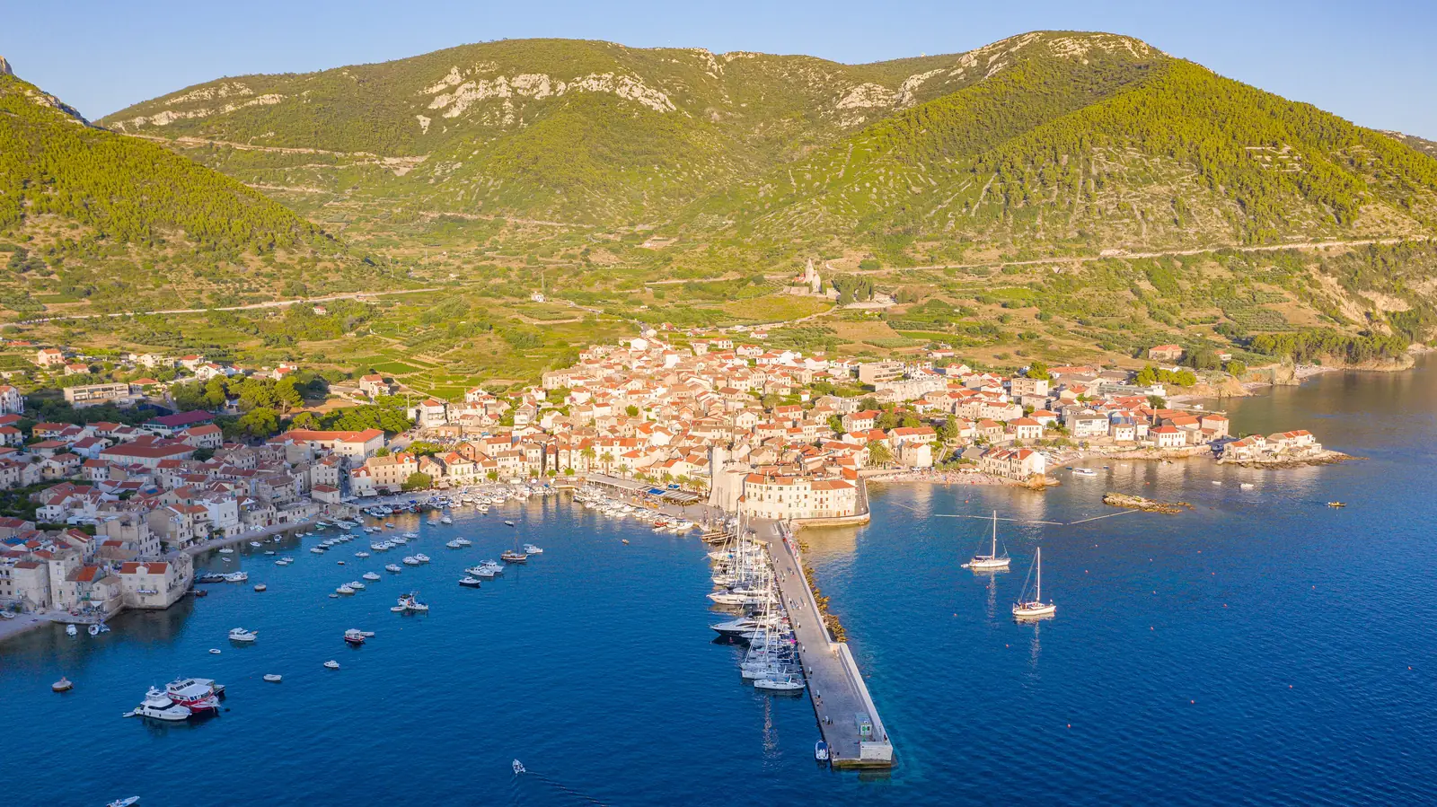 Aerial view of Komiza town and its harbor below green hills on Vis, Croatia