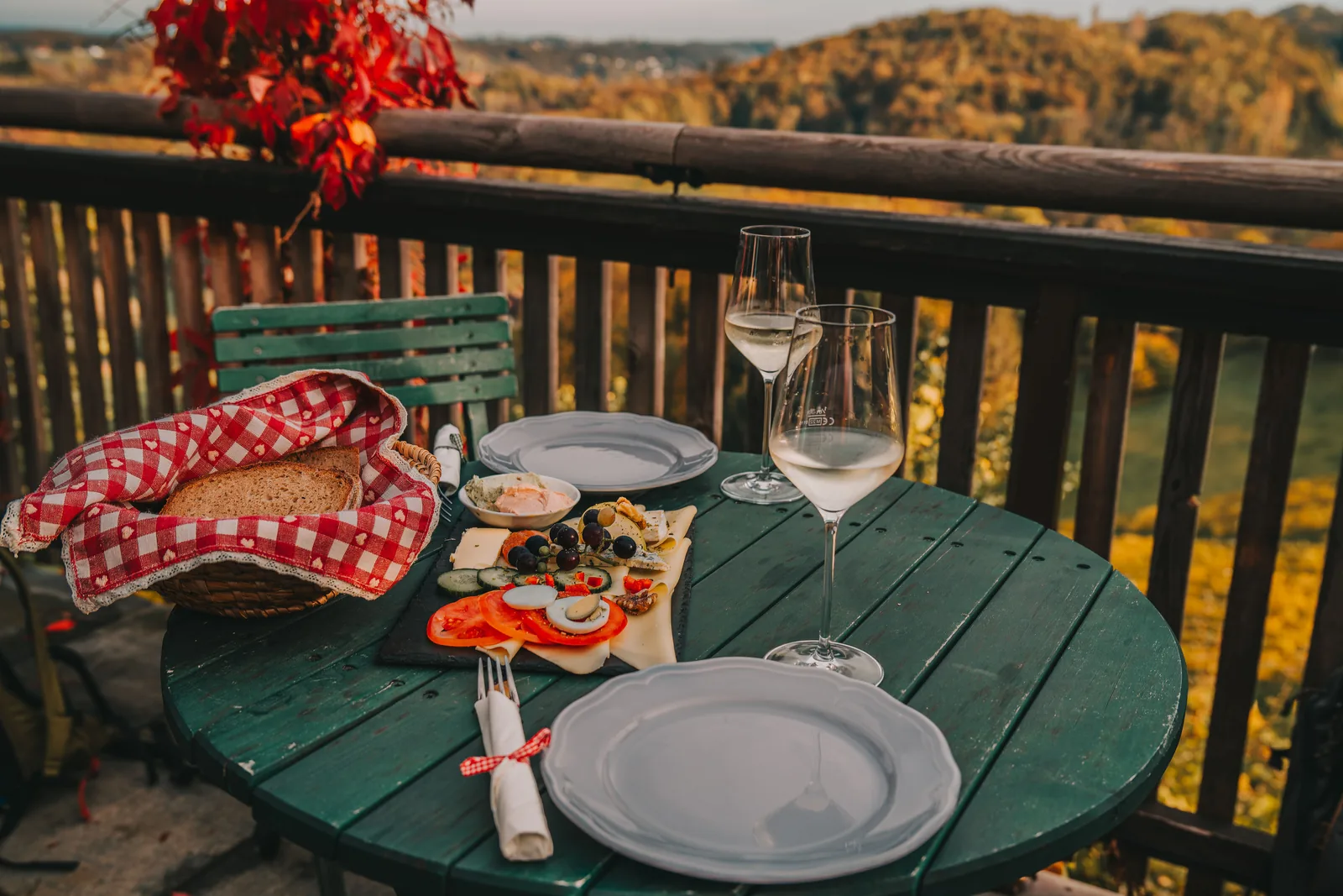 A wooden board of bread, cheese, and white wine on a terrace table overlooking the vineyards of Styria