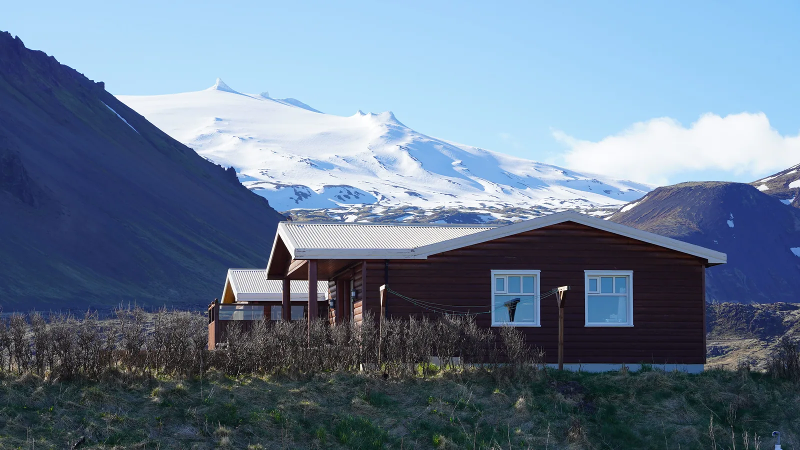 A dark red wooden cabin in the Icelandic countryside with a snow covered mountain behind