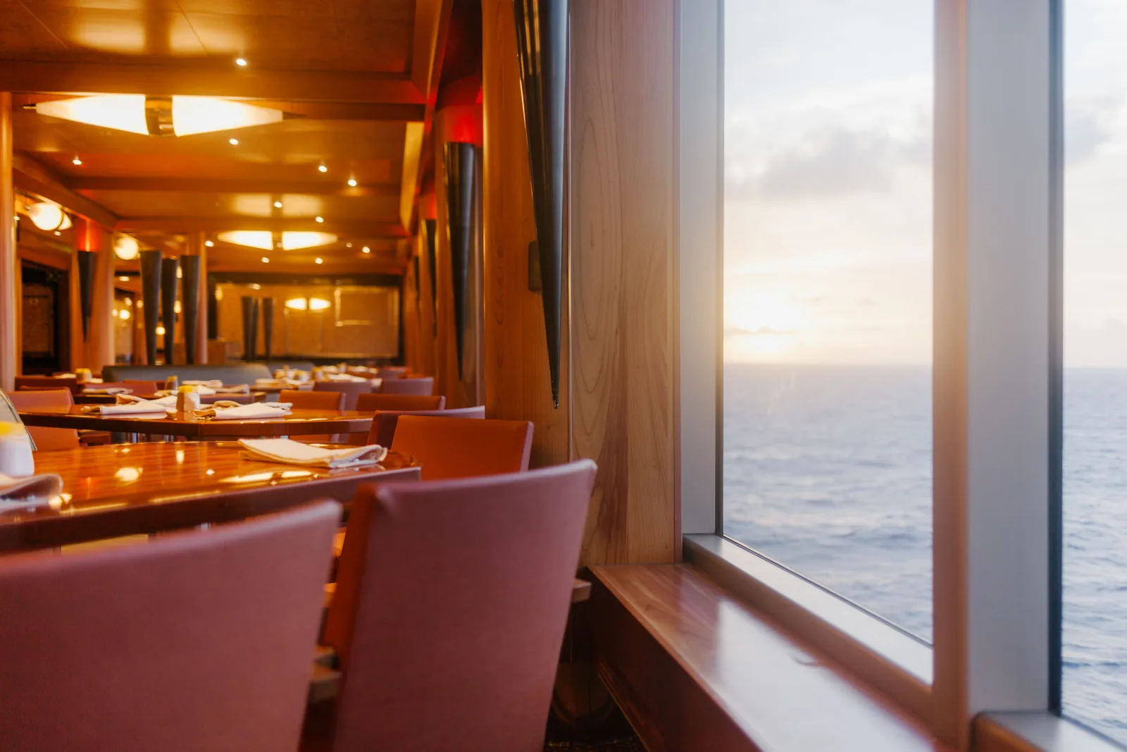 A cruise ship dining room with tables set along the windows at sunset