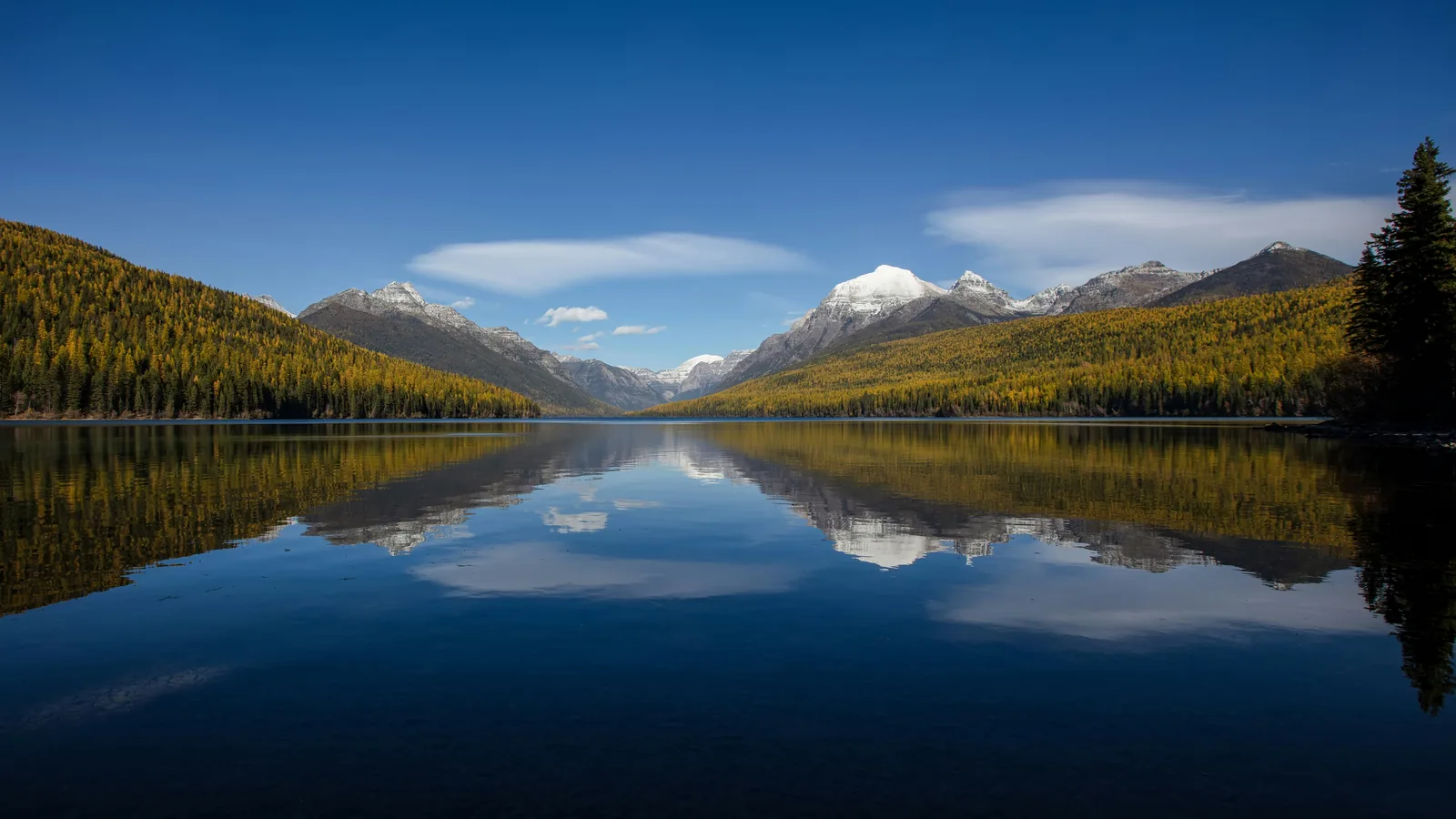 A long still lake with forested shores and distant peaks in Glacier National Park