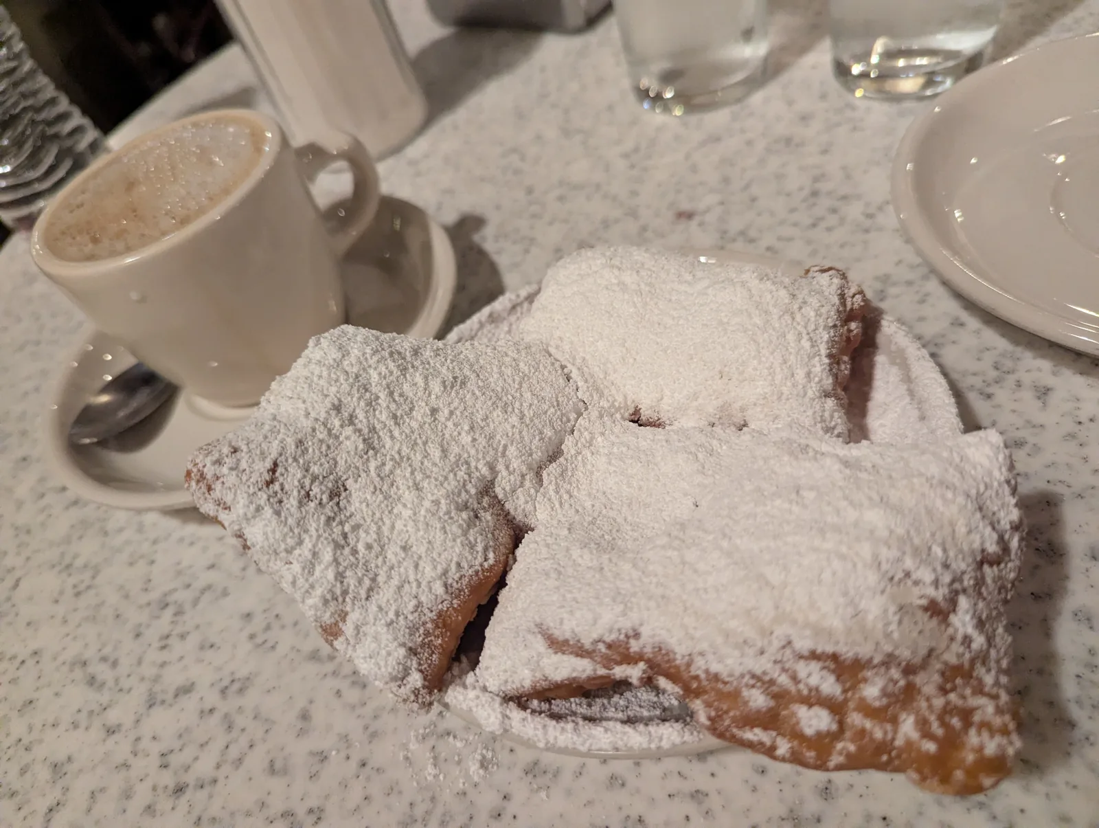 Three beignets buried in powdered sugar beside a cup of cafe au lait