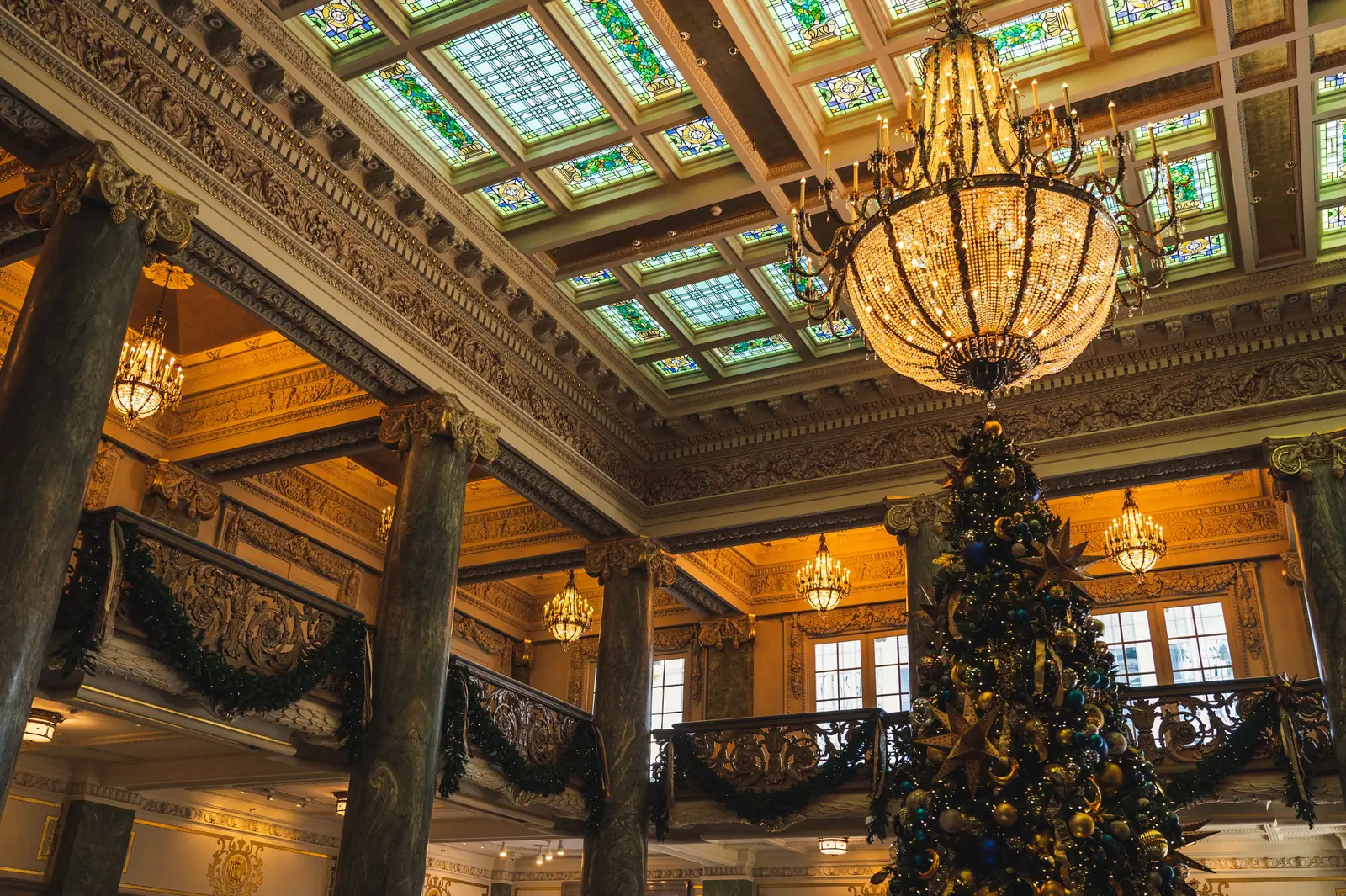A tall decorated Christmas tree standing under a chandelier in a grand hall