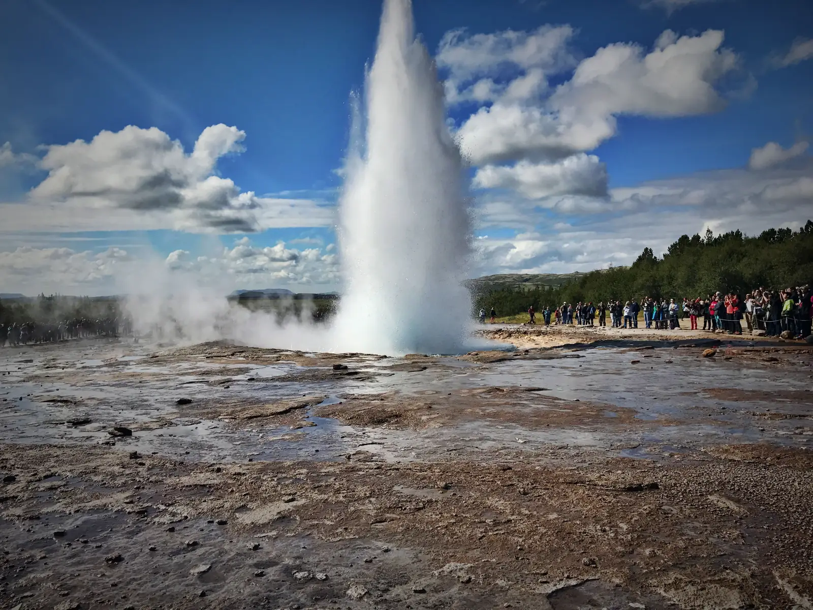 The Strokkur geyser erupting in a tall column of water while a crowd watches