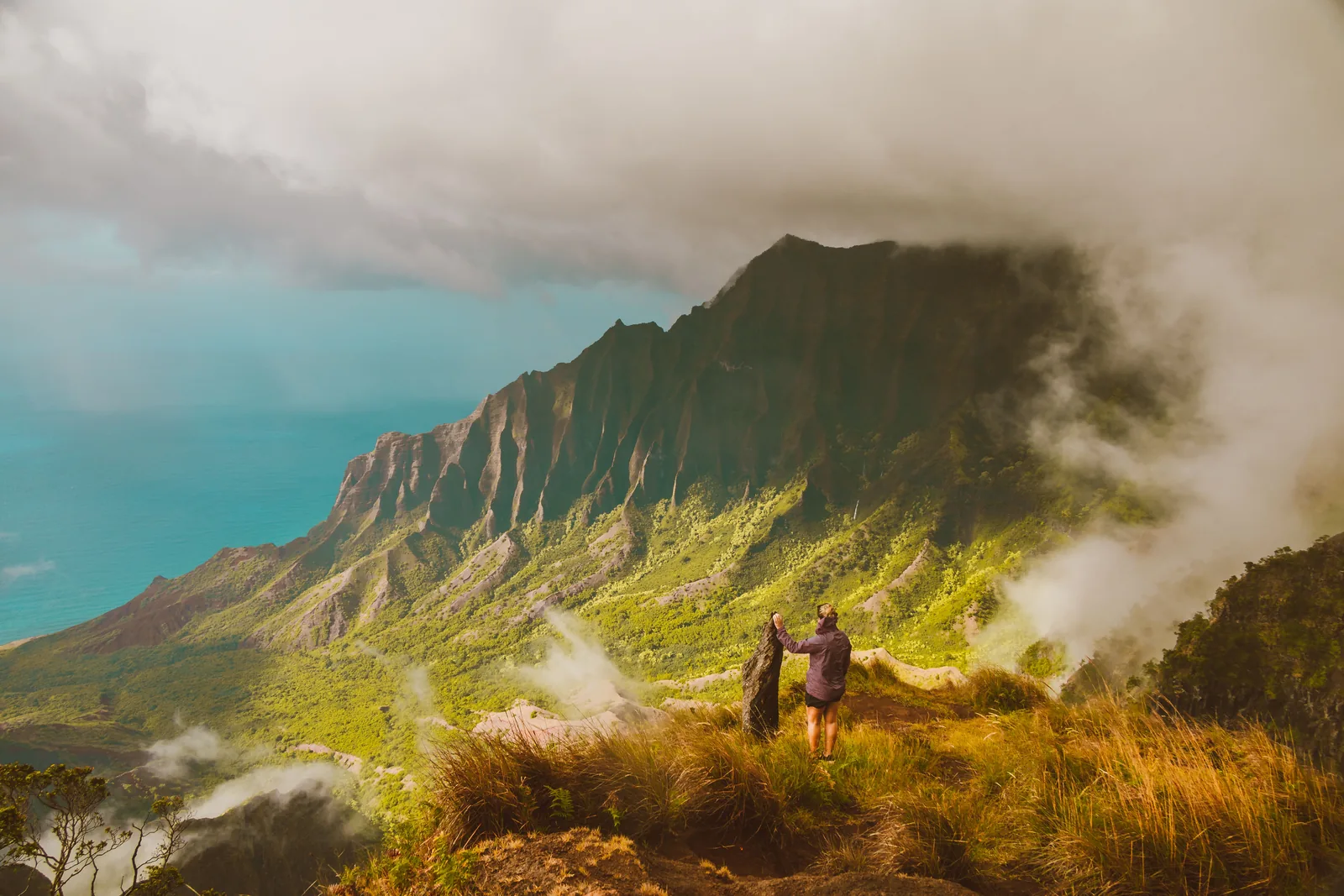 Two hikers standing on a grassy ridge above a misty valley on Kauai