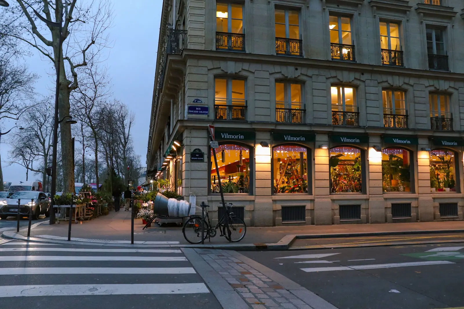 A Paris street corner at dusk in winter with lit shop windows and bare trees