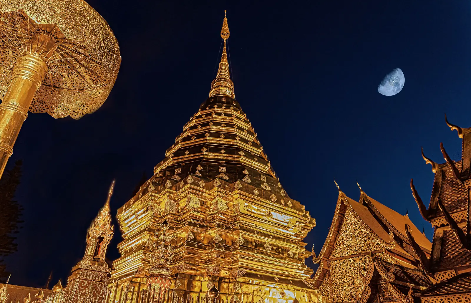 A golden temple spire lit at dusk with the moon above it in northern Thailand