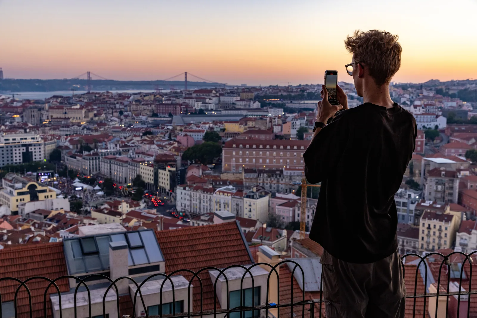 A person photographing the rooftops of Lisbon from a hillside viewpoint at sunset