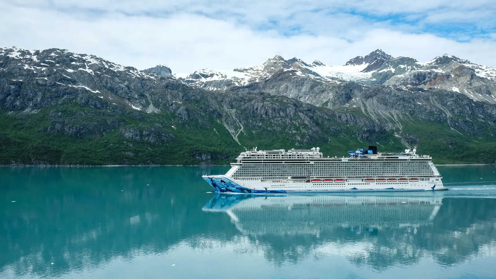 A cruise ship on calm green water below a glacier and snow covered peaks