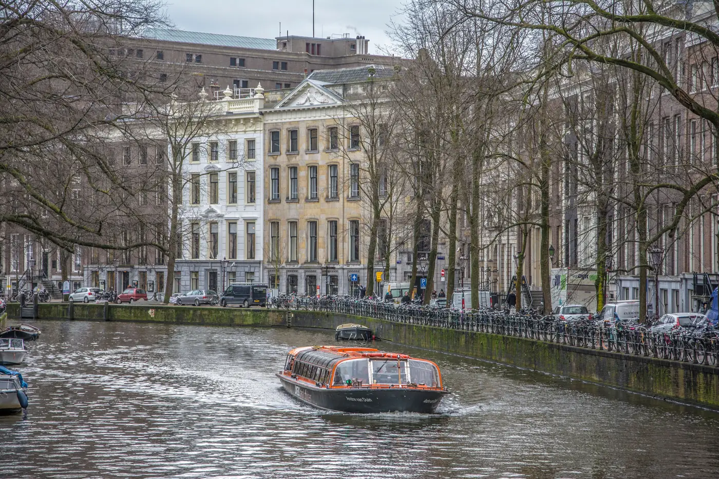 A glass topped canal cruise boat passing bare winter trees on an Amsterdam canal