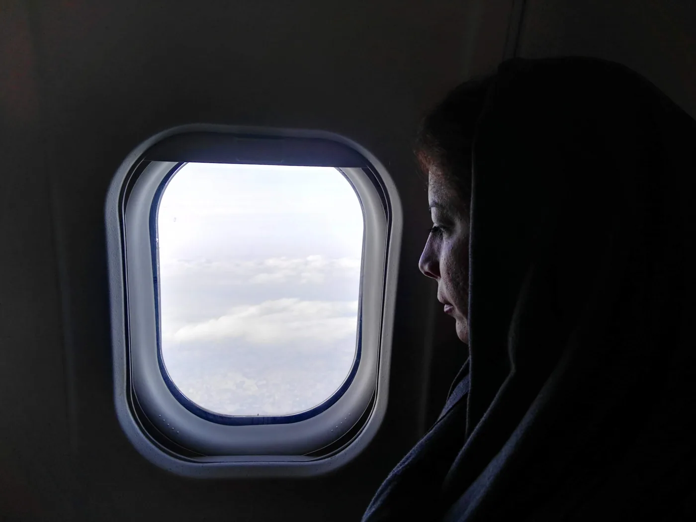 A traveler in a hood looking out an airplane window at the clouds