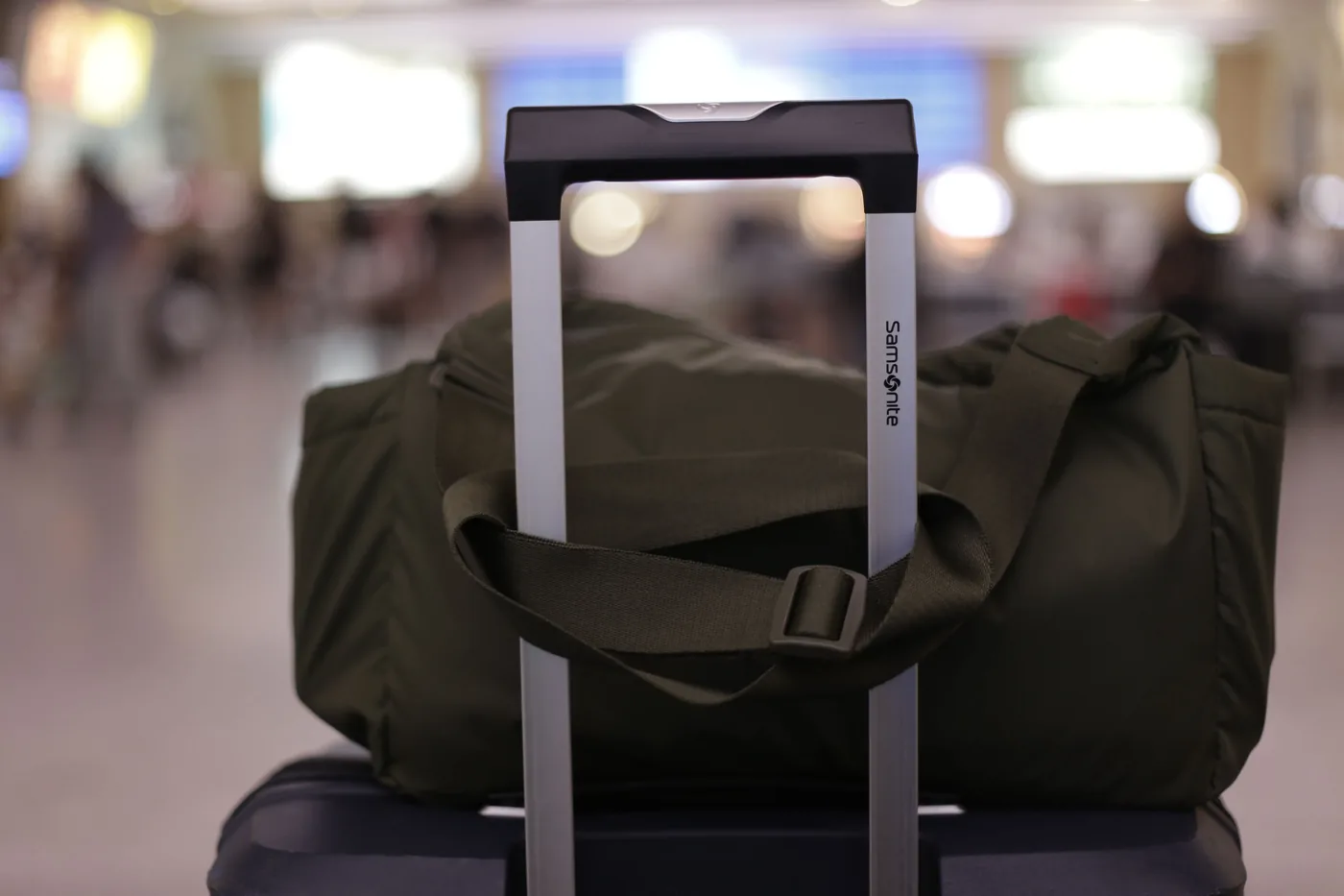 A green duffel bag sitting on a carry on suitcase handle in a busy airport terminal