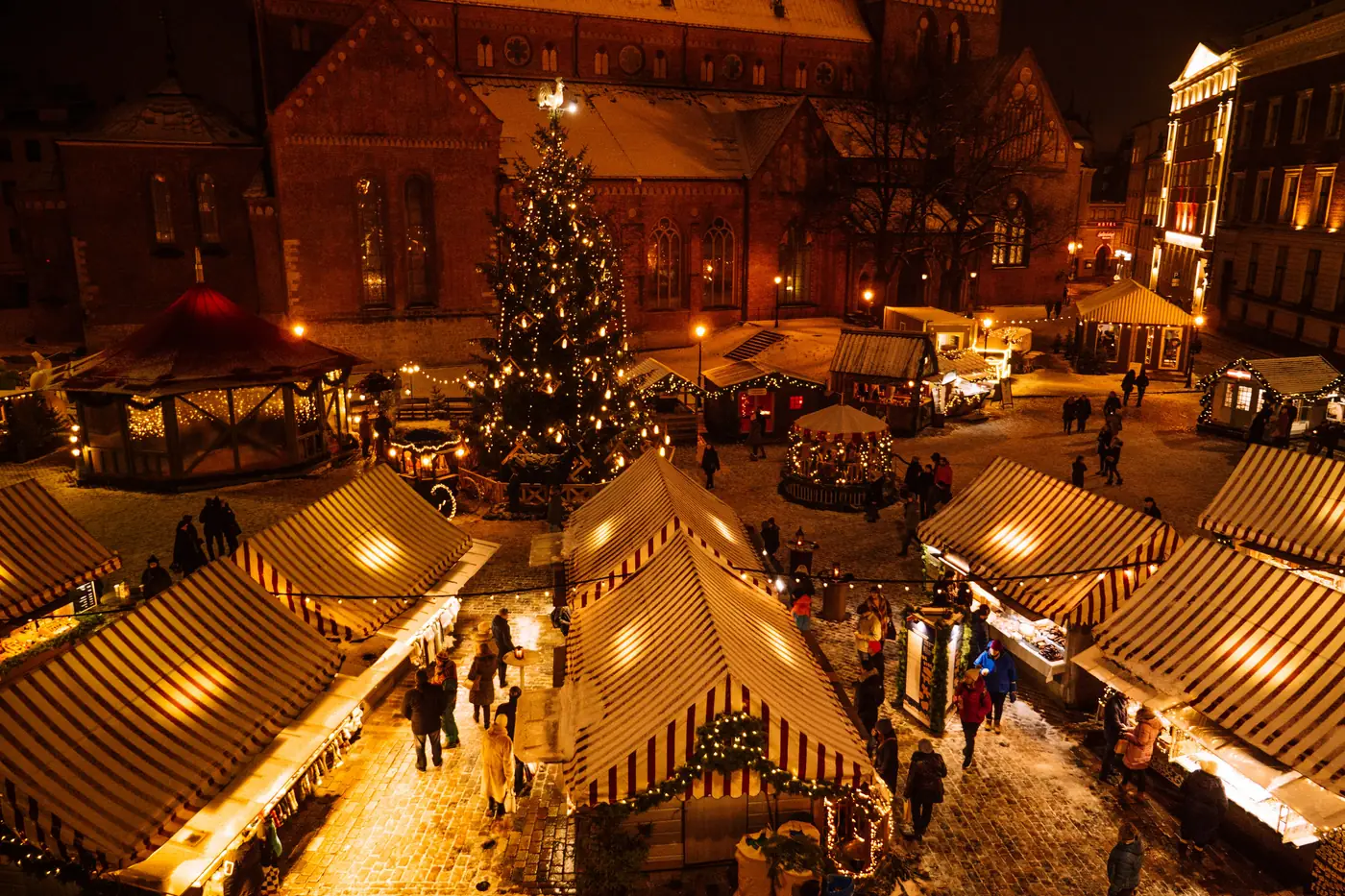 An overhead view of a snowy Christmas market lit up at night around a tall Christmas tree