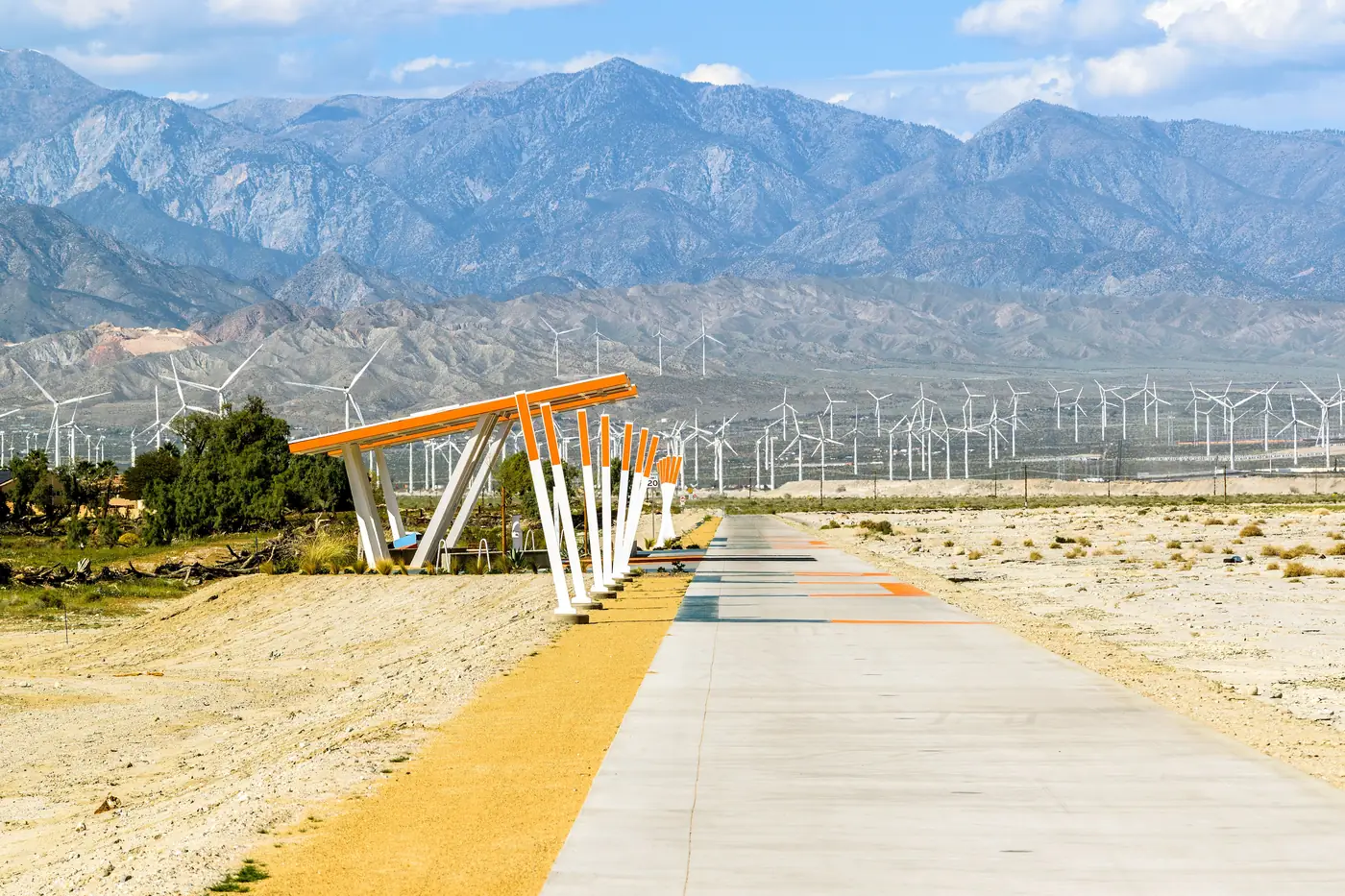 A paved desert path running toward the San Gorgonio wind farm with mountains behind