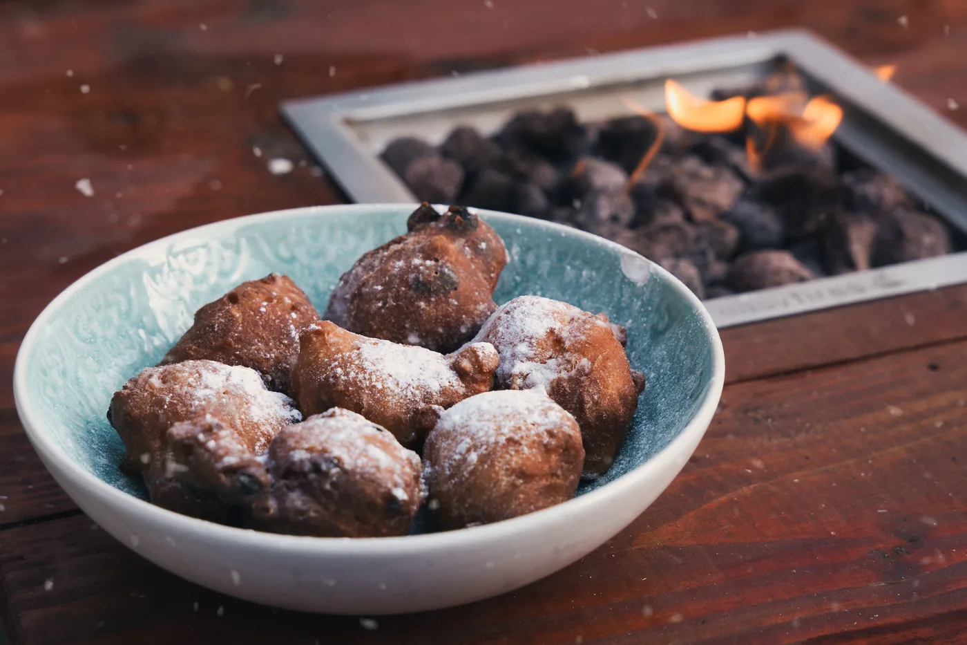 A bowl of Dutch oliebollen dusted with powdered sugar beside a warm fire