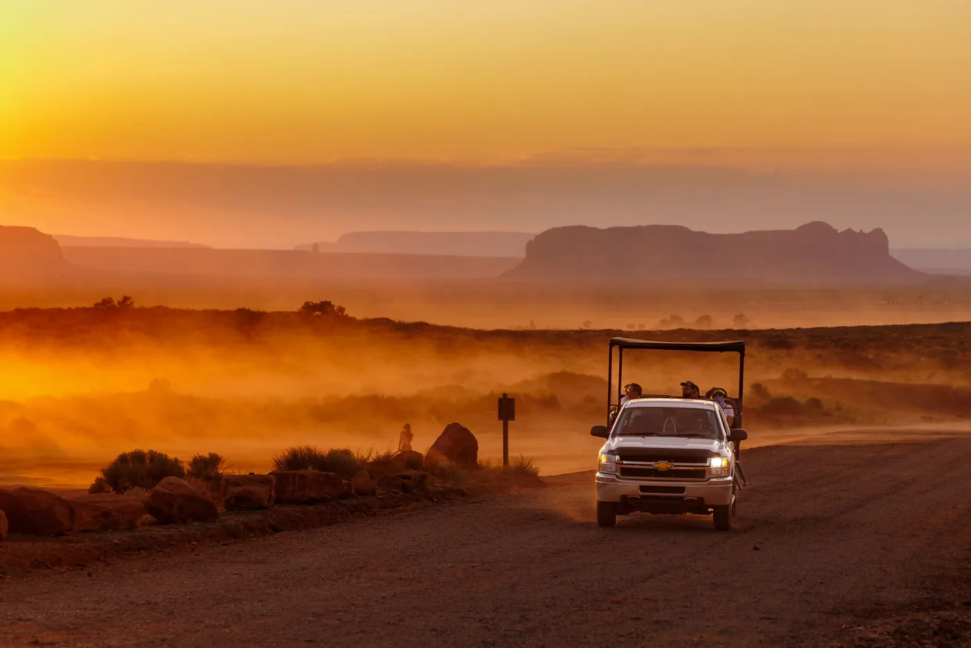 An open tour vehicle raising dust on a dirt road in Monument Valley at sunset