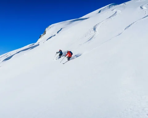 Two skiers descending a wide untracked snow slope under a deep blue sky