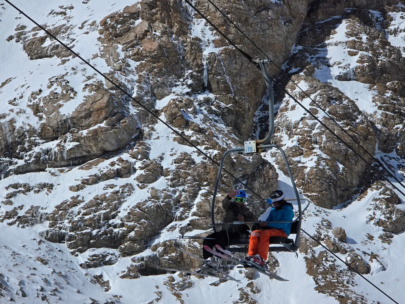 Two skiers in helmets and goggles riding a chairlift past a snowy rock face