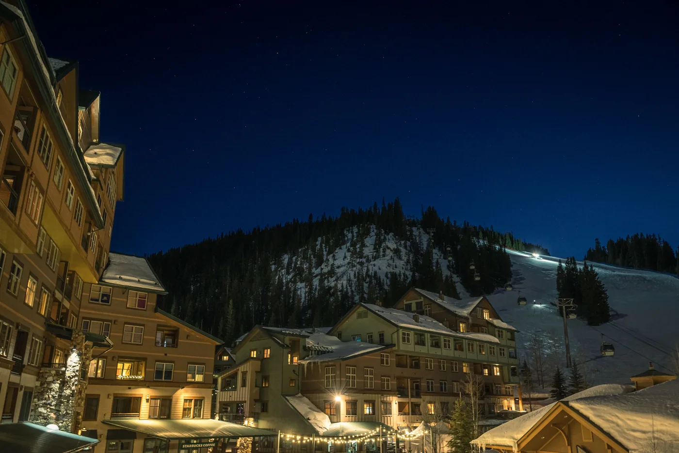 A lit ski village at dusk with a floodlit run on the mountain behind it