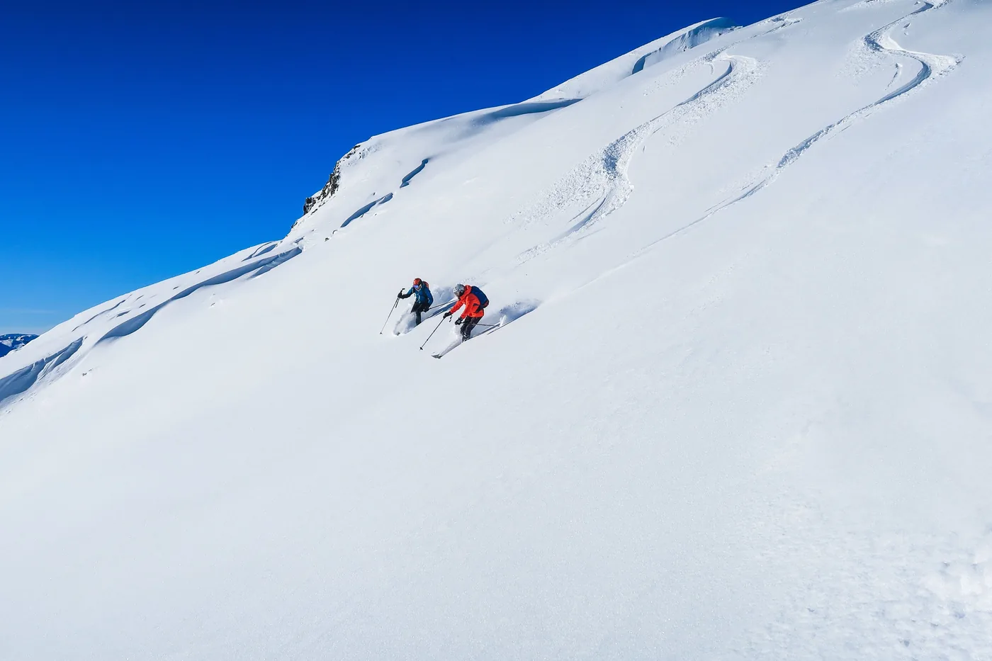 Two skiers descending a wide untracked snow slope under a deep blue sky