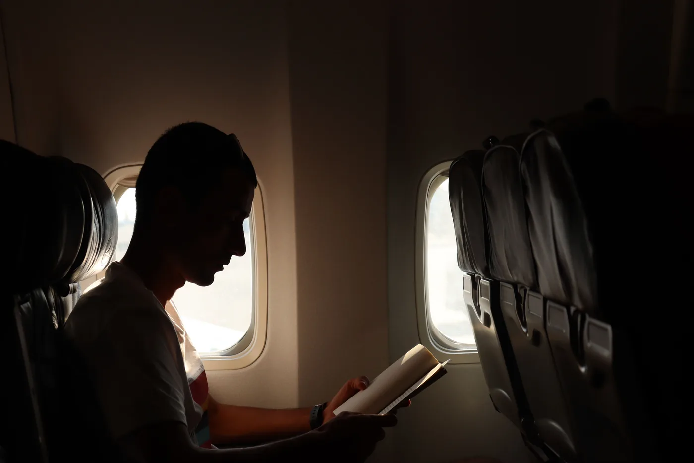 A passenger reading a book by the window in a dim airplane cabin