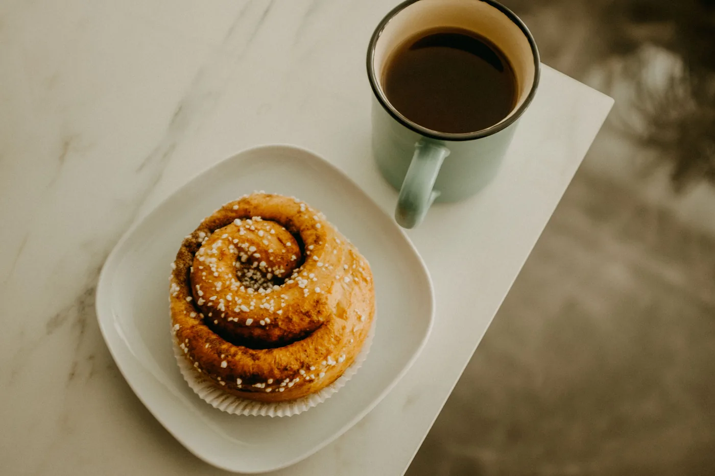 A cinnamon bun with pearl sugar beside a cup of coffee