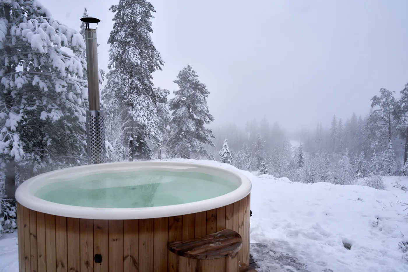 A wooden hot tub surrounded by snowy pine trees